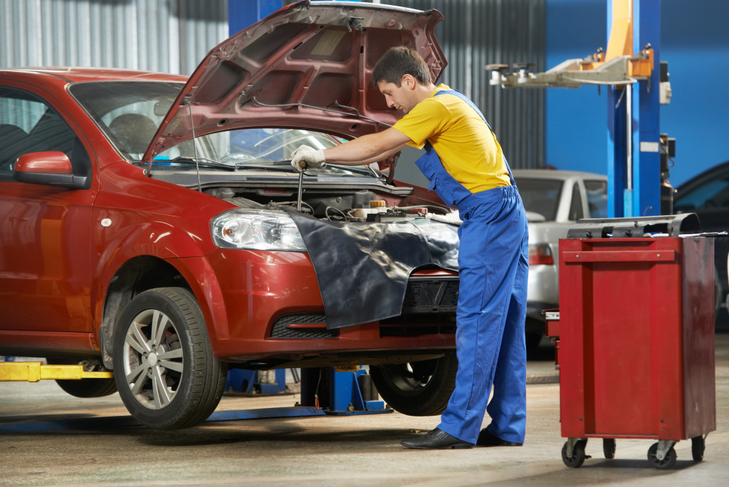 A mechanic in overalls is working under the hood of a car in an auto repair shop